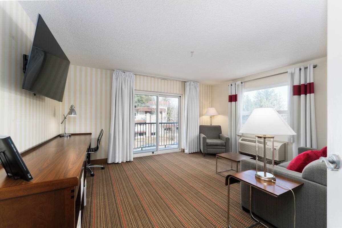 Ein Hotelzimmer mit einem Holztisch, einer Couch, einem an der Wand befestigten Fernseher, einer Lampe und Blick ins Freie.
