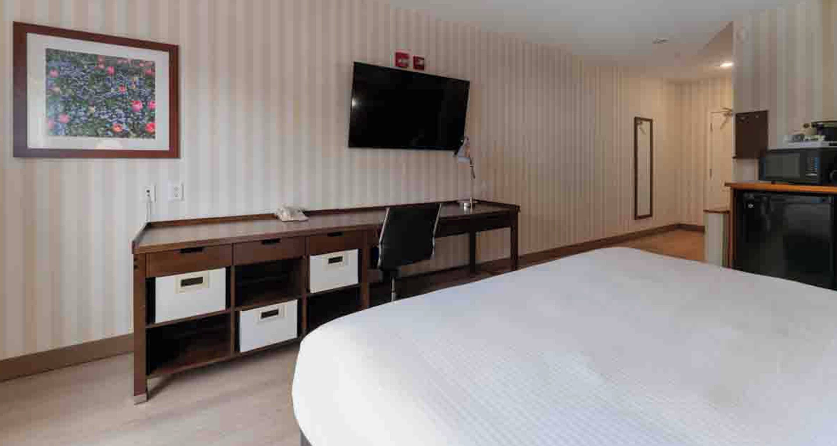A hotel room with a bed, a desk with drawers, a chair, a television mounted on the wall, and a microwave on a counter.