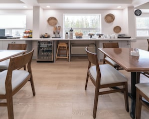 Eine moderne Küche mit Holzkstühlen, einem Kühlschrank, einer Kaffeemaschine und einem Fenster mit Blick auf Bäume.