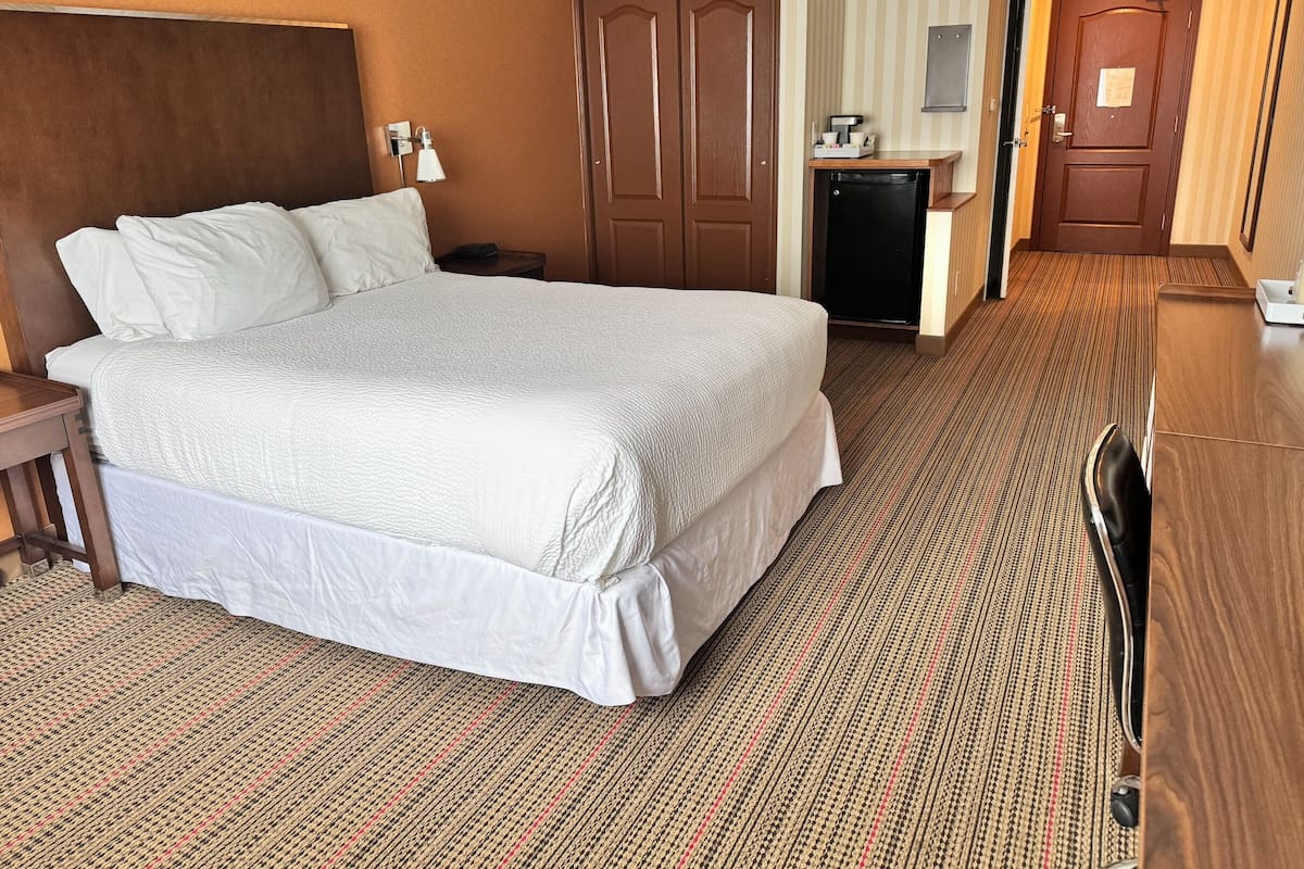 Ein Hotelzimmer mit einem großen Bett, einem Schreibtisch mit Computer, einem Kleiderschrank und einer Tür.