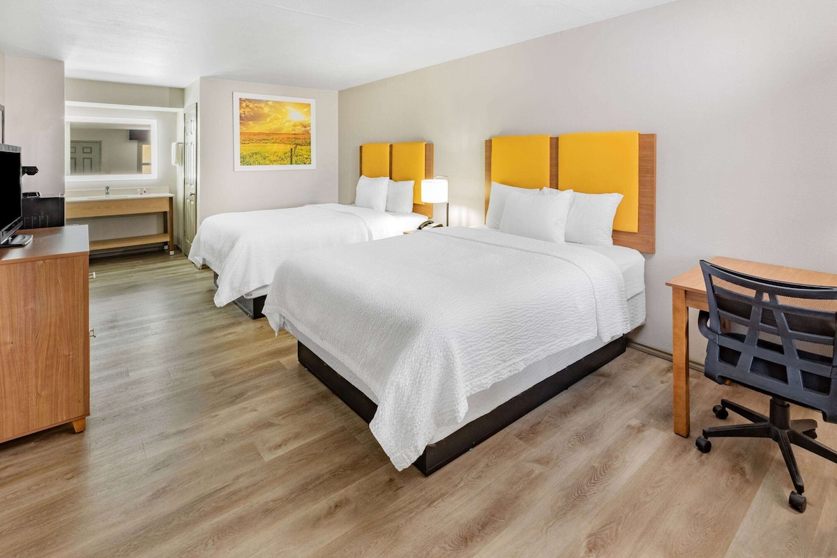 A hotel room with two beds, a desk, a chair, a TV, and a bathroom with a mirror and sink.