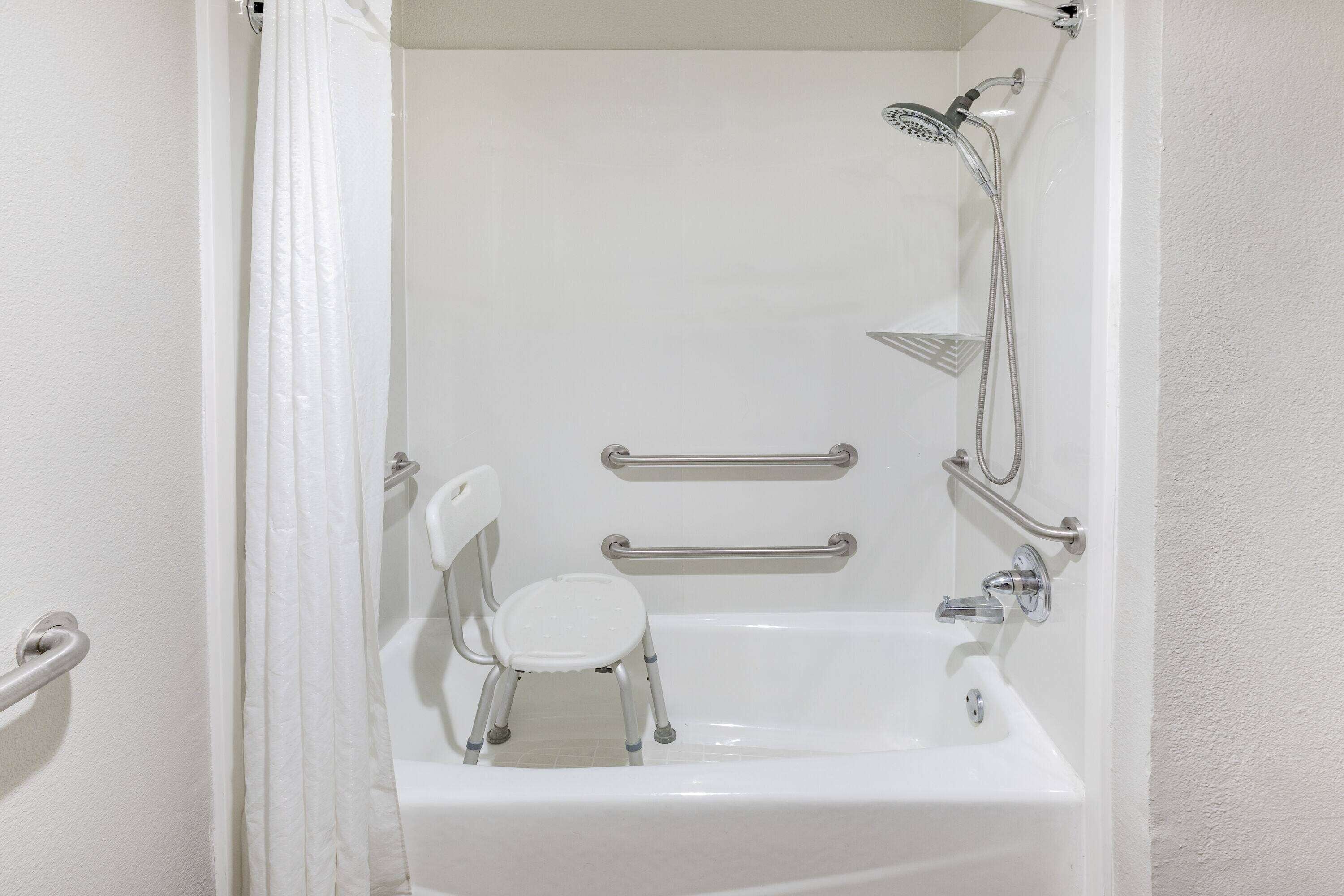 A bathroom with a white bathtub, a small white stool, and a shower with grab bars.