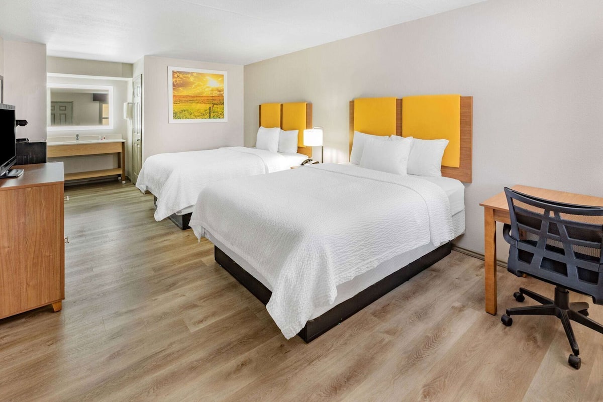A hotel room with two beds, a desk, a chair, a TV, and a bathroom visible in the background.