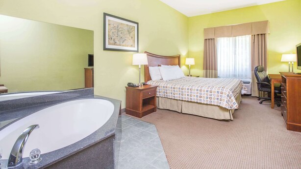 A hotel room with a large bathtub, a bed with a patterned bedspread, a desk with a chair, and a desk lamp. - Days Inn & Suites by Wyndham Swainsboro (Swainsboro)