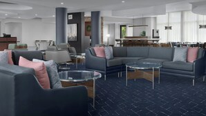 Un hall d’hôtel moderne avec un tapis bleu foncé, des canapés gris agrémentés de coussins roses et gris, une table basse en verre et un comptoir d’accueil doté d’un ordinateur et d’une plante.