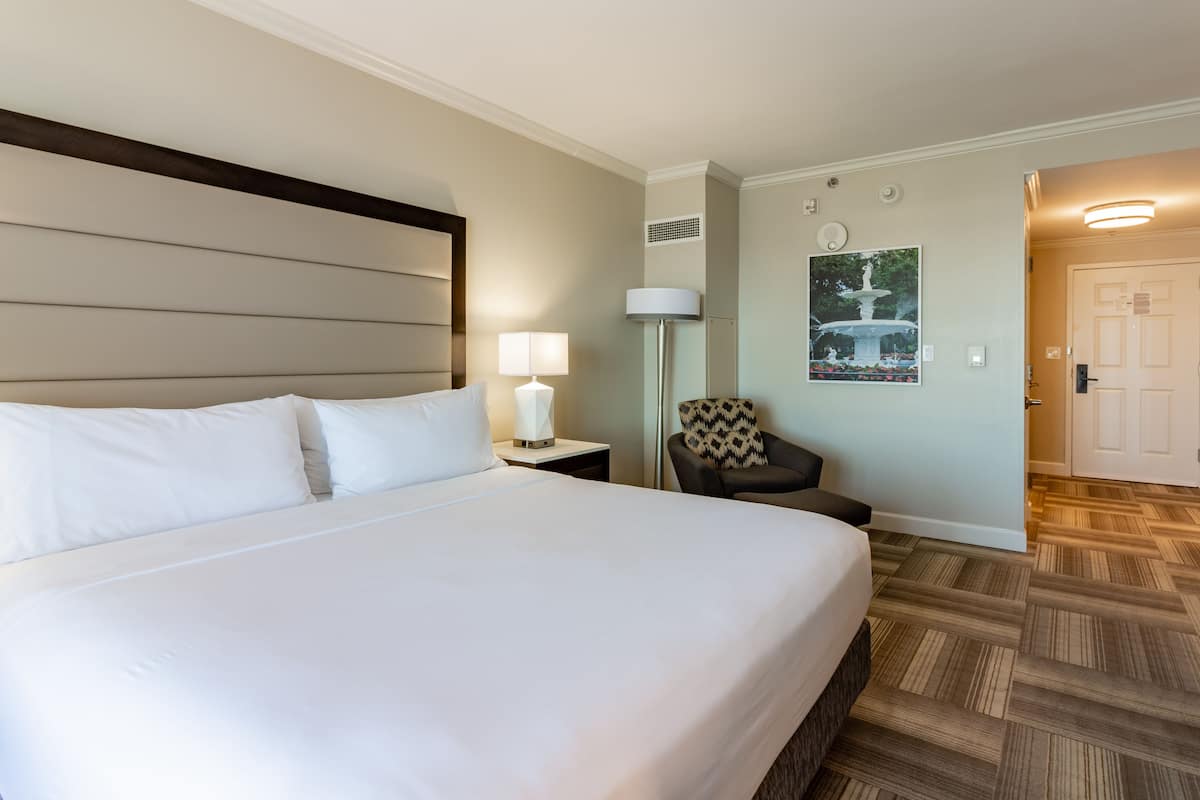 A hotel room with a large bed, a nightstand with a lamp, a chair, a poster on the wall, and a door.