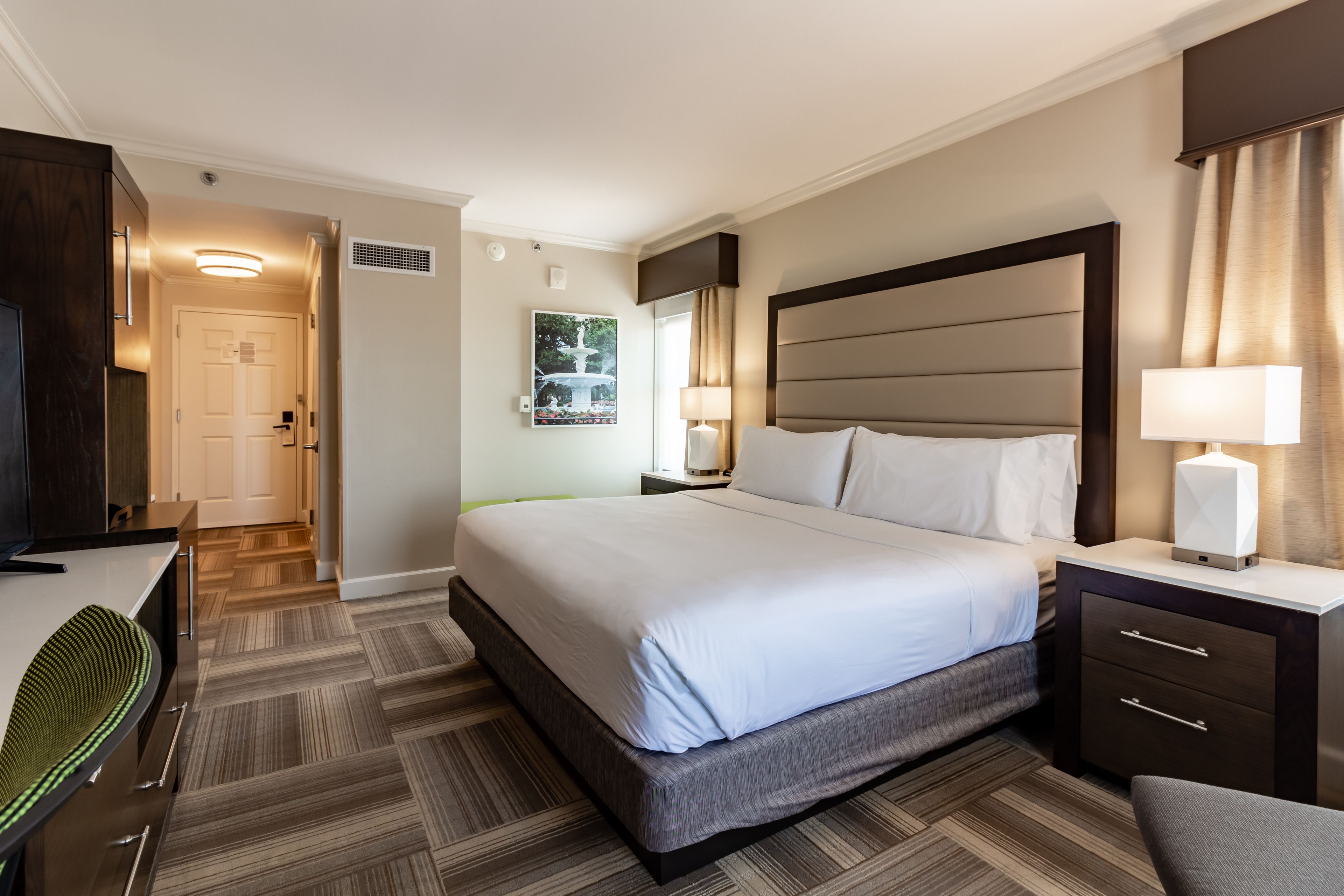 A hotel room with a large bed, a desk, a chair, and a nightstand with a lamp.