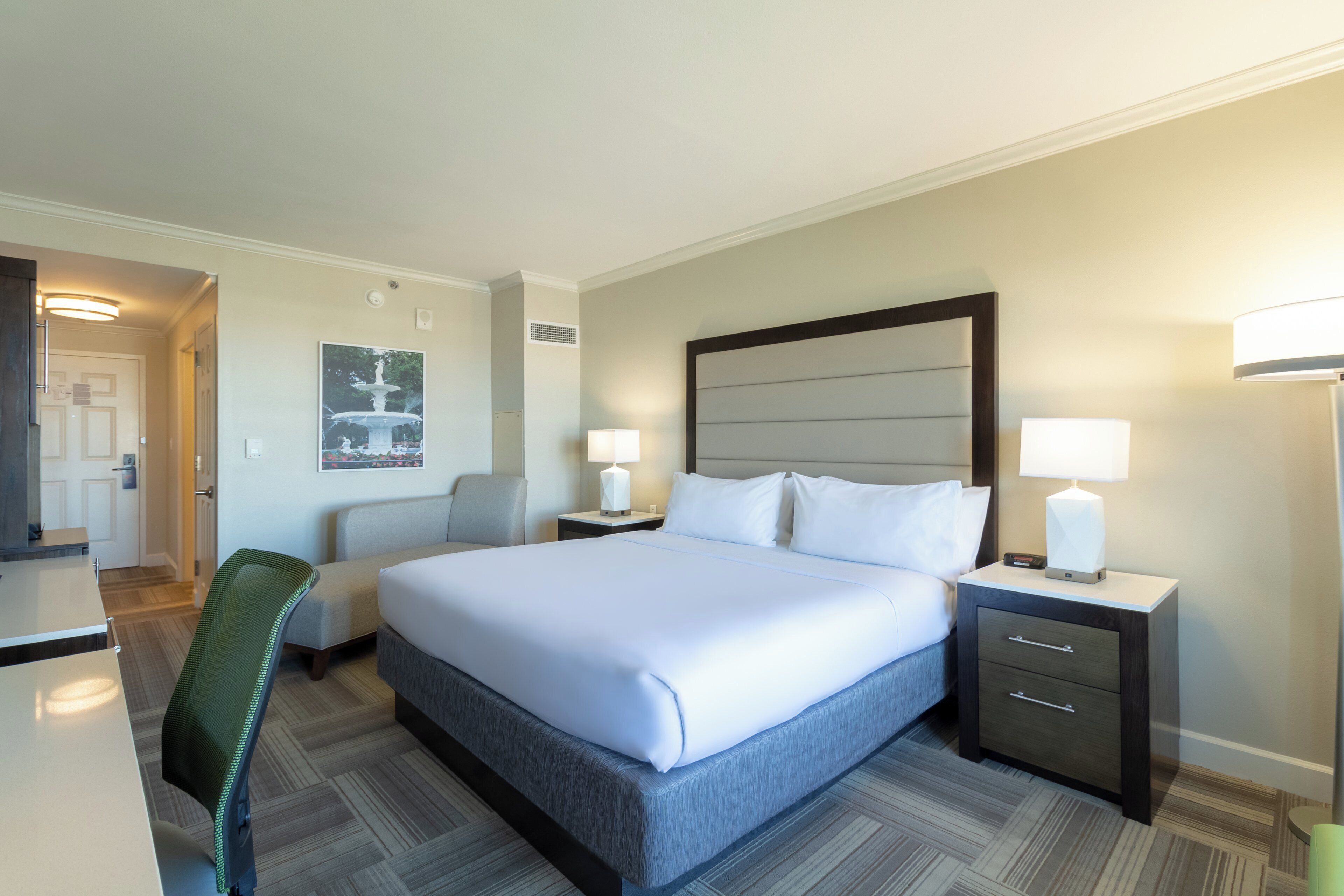 A hotel room with a bed, two bedside tables with lamps, a chair, a desk, and a painting on the wall.