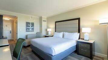 A hotel room with a bed, two bedside tables with lamps, a chair, a desk, and a painting on the wall.