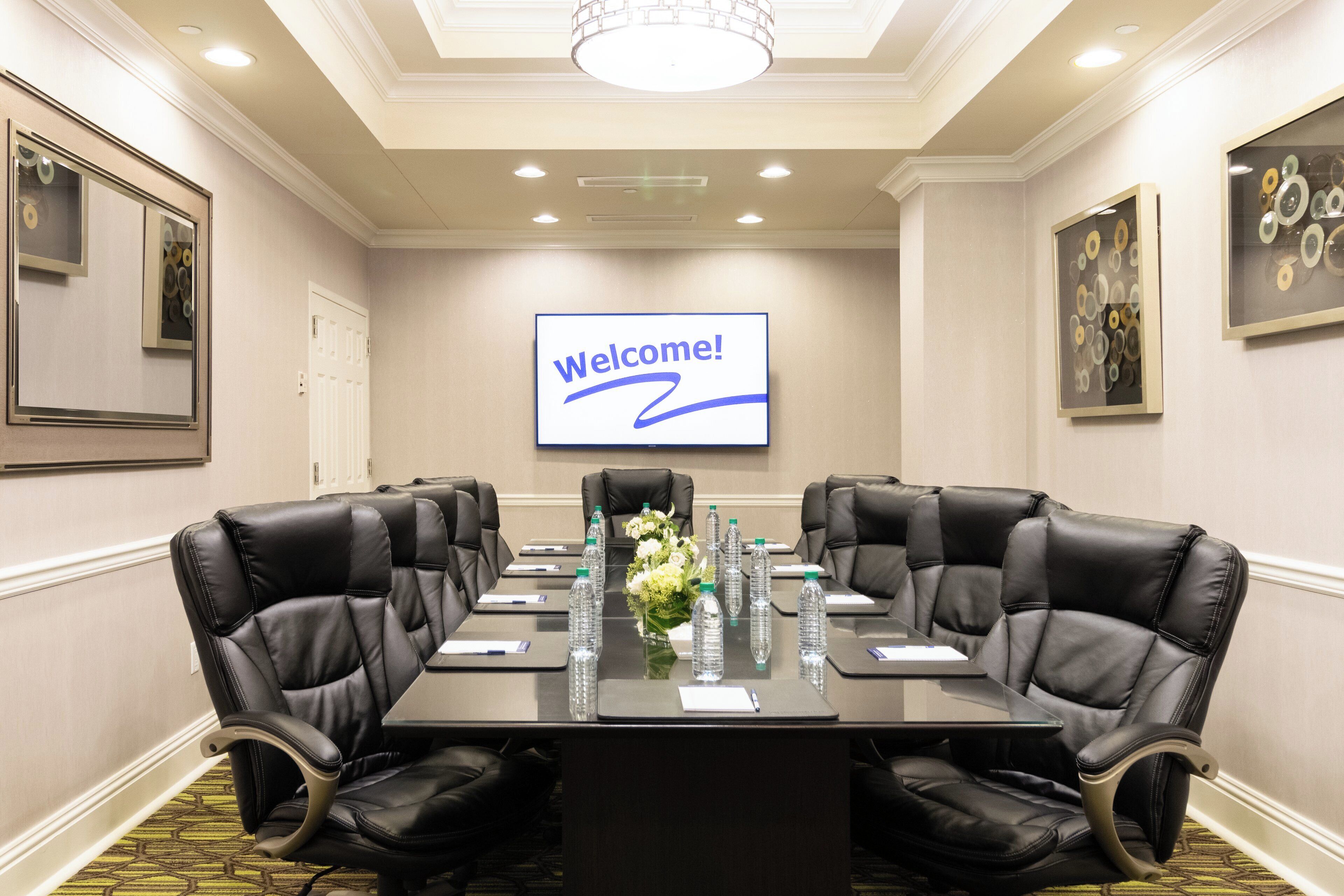 Un salón de conferencias con un cartel que dice '¡Bienvenido!', sillas de cuero, una larga mesa con botellas de agua y un arreglo floral.