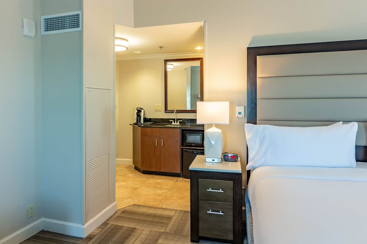 A hotel room with a bed, bedside table, a mirror, a small kitchenette with a microwave, and a coffee maker.