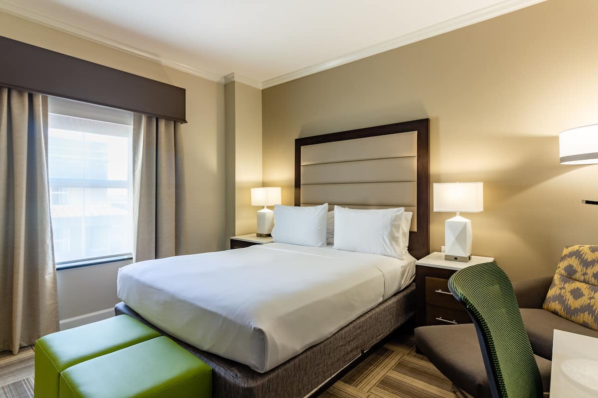 A hotel room with a large bed, two bedside lamps, a green ottoman, a chair, and a view of the city through the window.