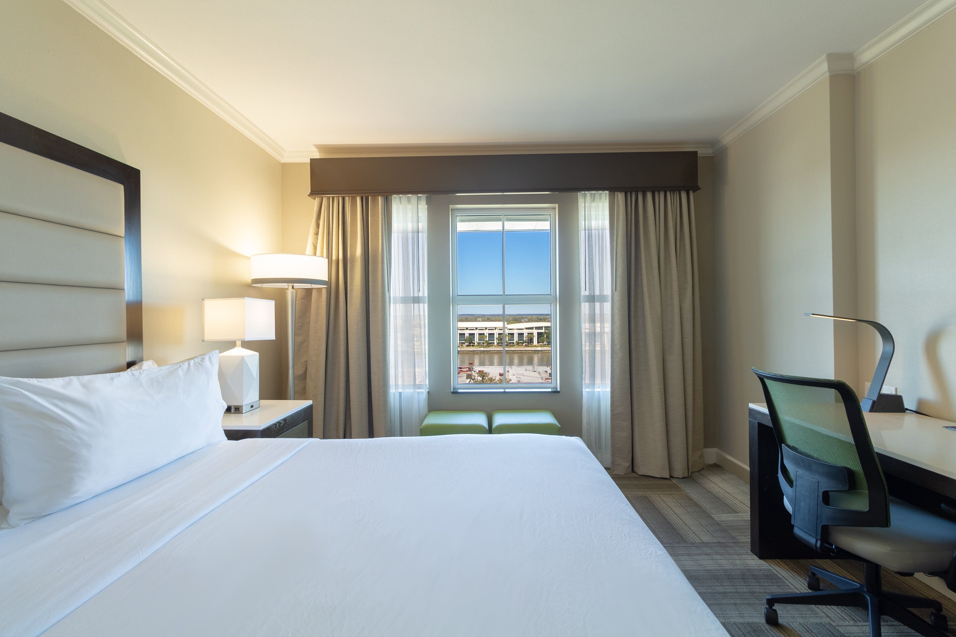 A hotel room with a large bed, a desk, a chair, a lamp, and a view of a building through a window.