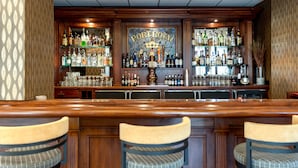 Een bar met een houten toonbank, drie barkrukken en verschillende flessen en glazen.