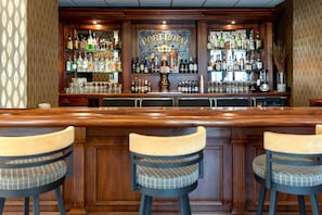 A bar area with a wooden counter, three bar stools, and a variety of bottles and glasses.
