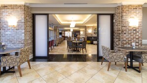 Een ruime hotellobby met een modern design, met een centraal gelegen eetgedeelte met ronde tafels en stoelen, en accentmuren van baksteen.