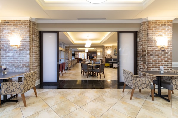 A spacious hotel lobby with a modern design, featuring a central dining area with round tables and chairs, and brick accent walls.