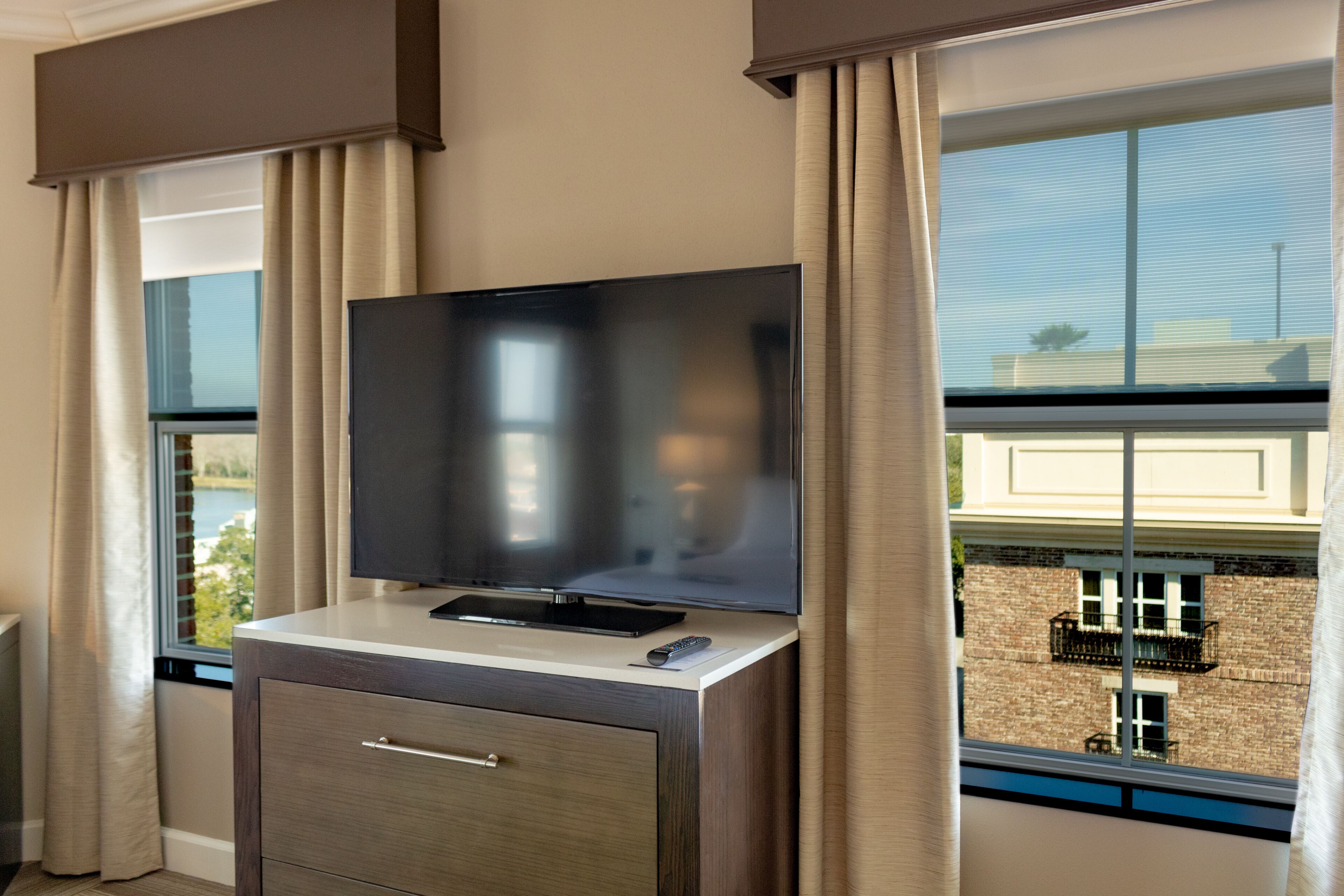 Ein Hotelzimmer mit einem Flachbildfernseher auf einer Holzkabine, einer Fernbedienung und einem Fenster mit Blick auf ein Gebäude und Bäume.