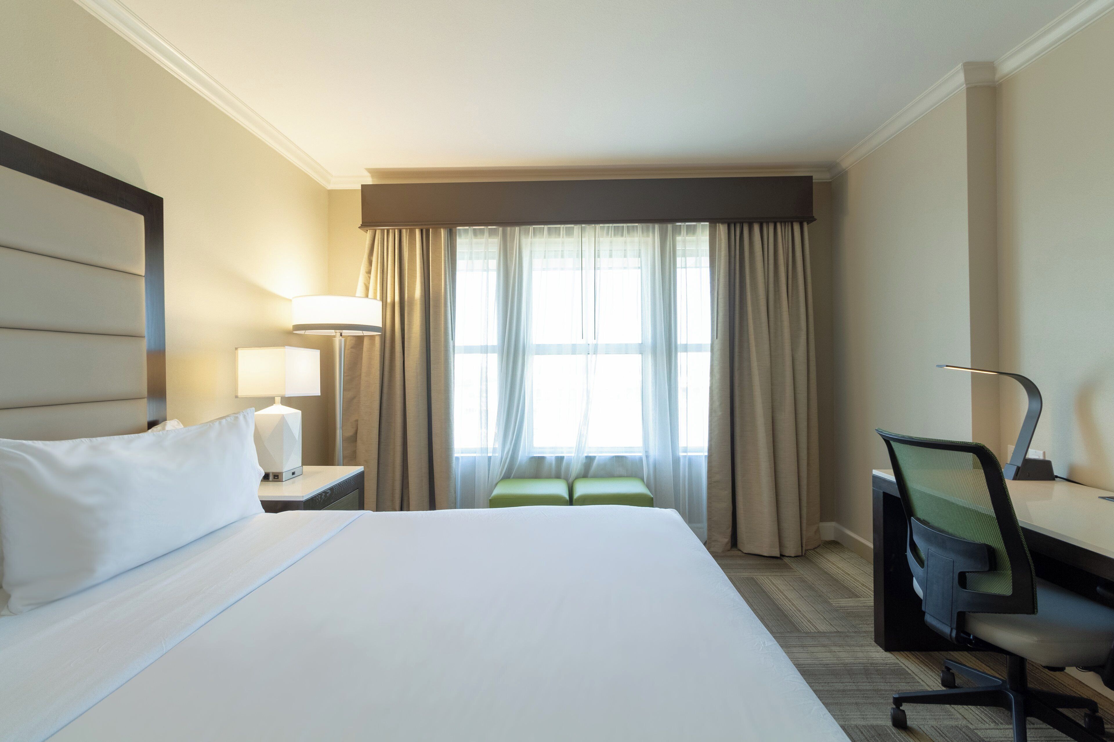 Una habitación de hotel con una cama grande, un escritorio, una silla y un ventanal con cortinas.