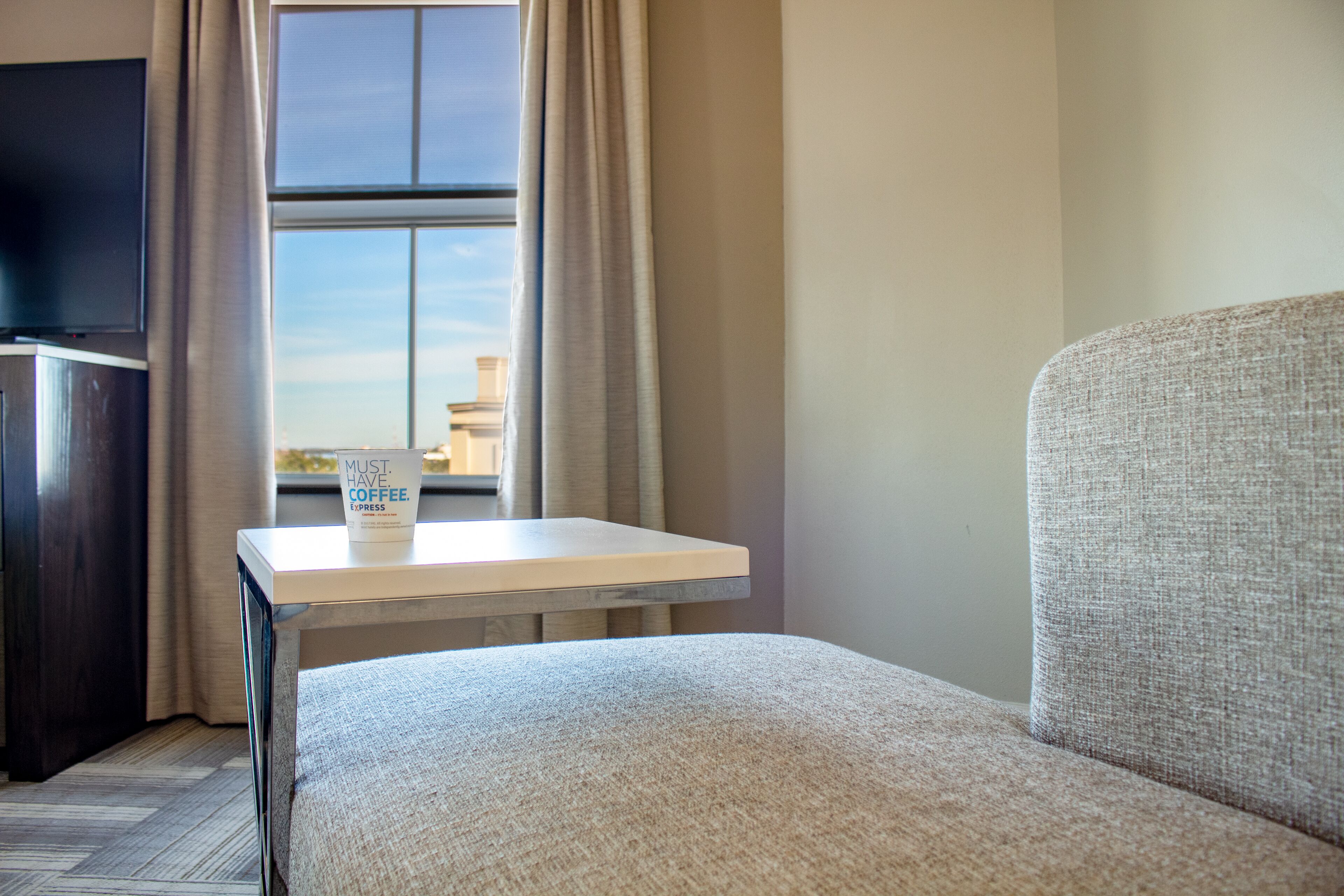 A hotel room with a sofa, a small table with a coffee cup, a TV, and a window with curtains.