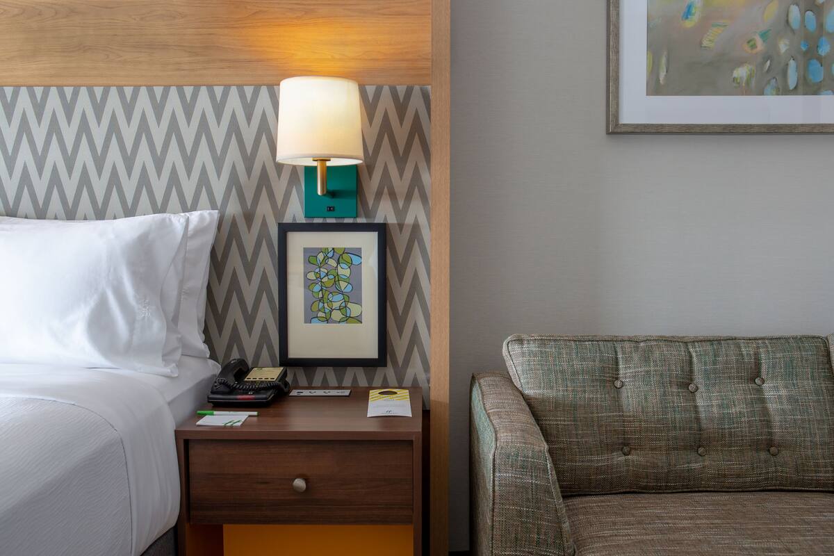 A hotel room with a bed, a nightstand with a lamp, a framed picture, and a couch.