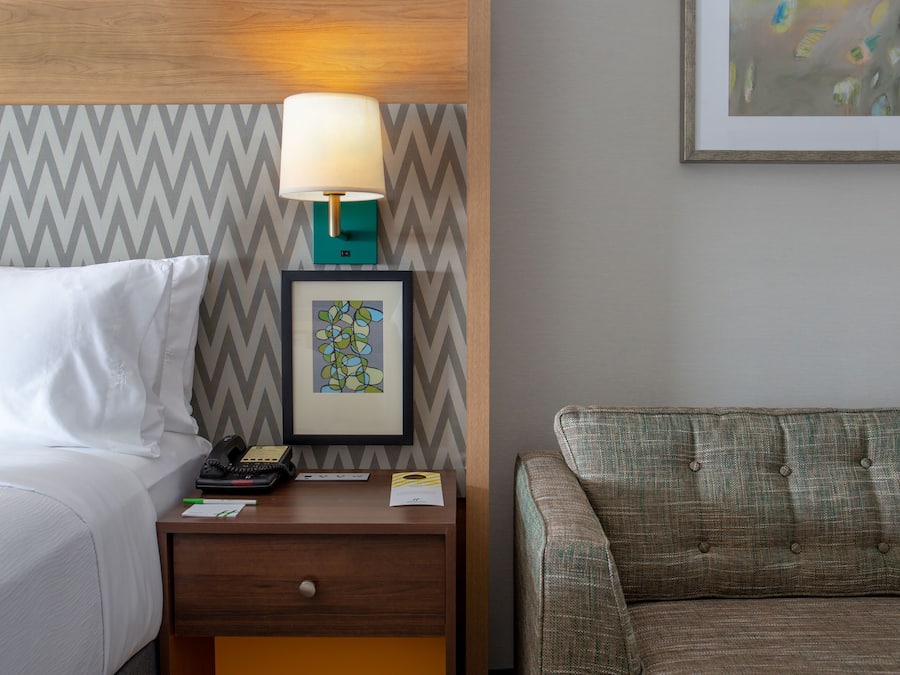 Habitación de hotel con cama, mesita de noche con lámpara, un cuadro enmarcado y un sofá.