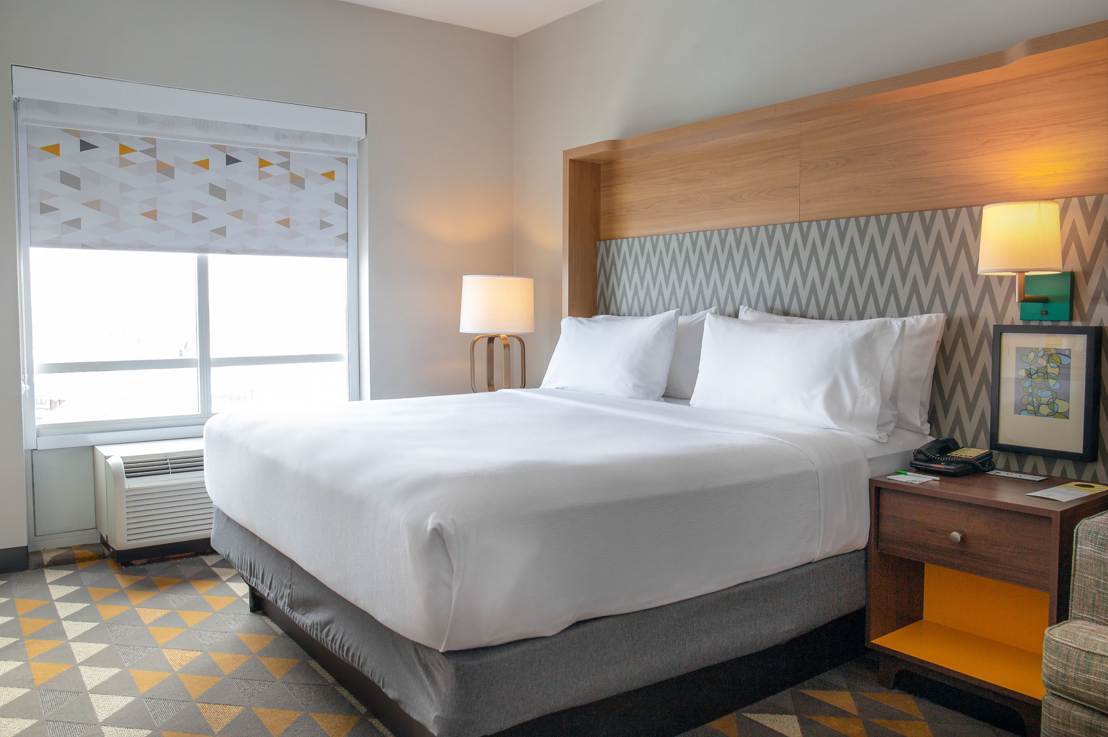 A hotel room with a large bed, a nightstand with a lamp, a framed picture, and a window with a geometric pattern.