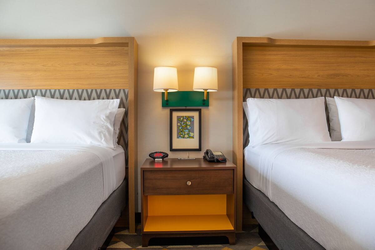 A hotel room with two beds, a nightstand with a lamp, a phone, and a framed picture.