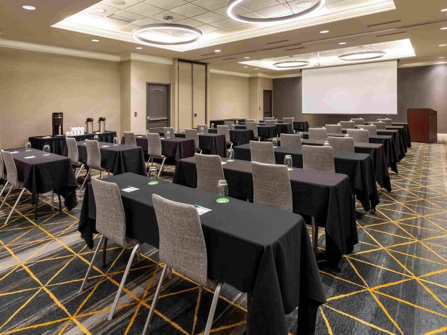 Una sala conferenze con file di tavoli e sedie, dotata di macchinette da caffè e bottiglie d'acqua, pavimento rivestito di moquette e schermo per proiettore.