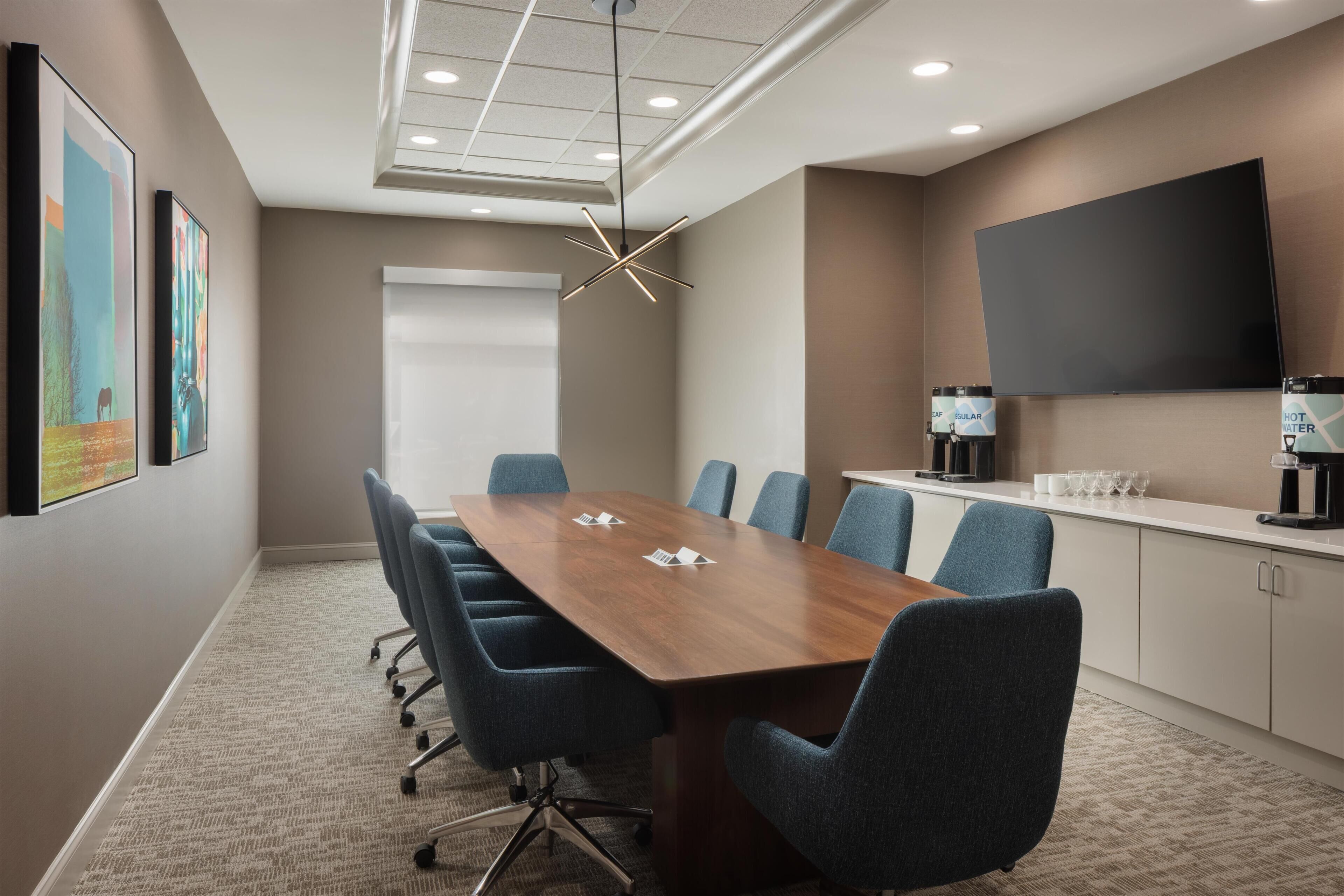 A conference room with a long table, blue chairs, a mounted TV, and artwork on the walls.