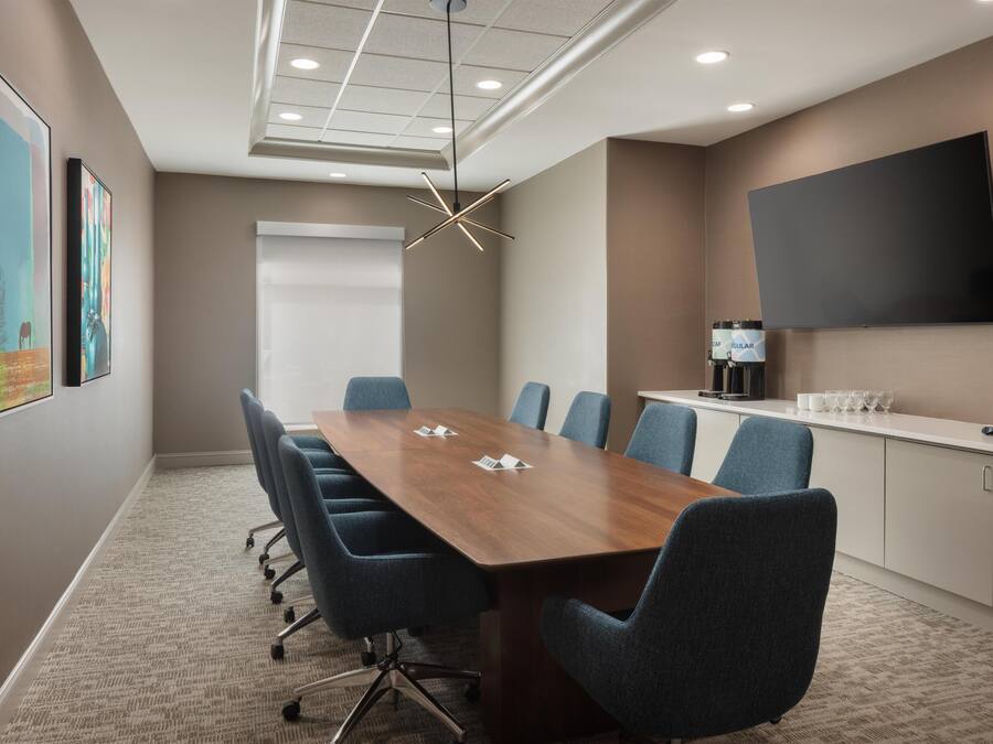 A conference room with a long table, blue chairs, a mounted TV, and artwork on the walls.
