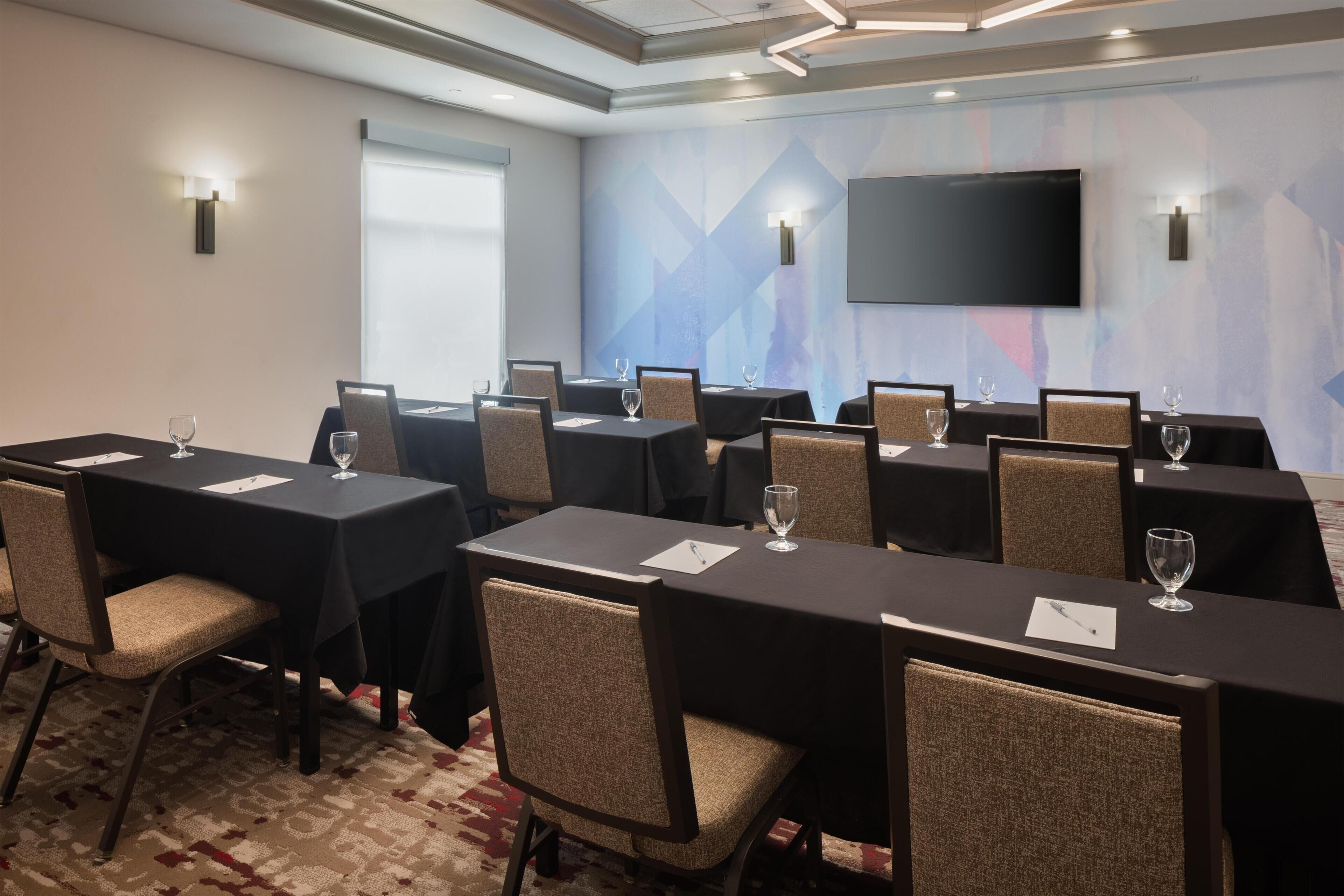A conference room with multiple tables, chairs, and a large flat-screen TV on the wall.