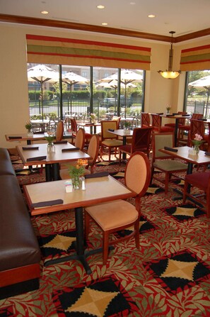 A restaurant with tables and chairs, a patterned carpet, and large windows with umbrellas outside. - Hilton Garden Inn Bowling Green (Bowling Green)