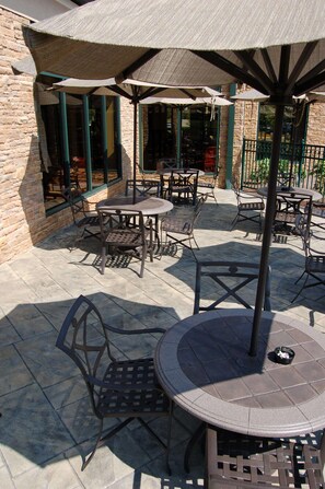 An outdoor patio area with multiple tables and chairs, shaded by umbrellas, and a stone building in the background. - Hilton Garden Inn Bowling Green (Bowling Green)