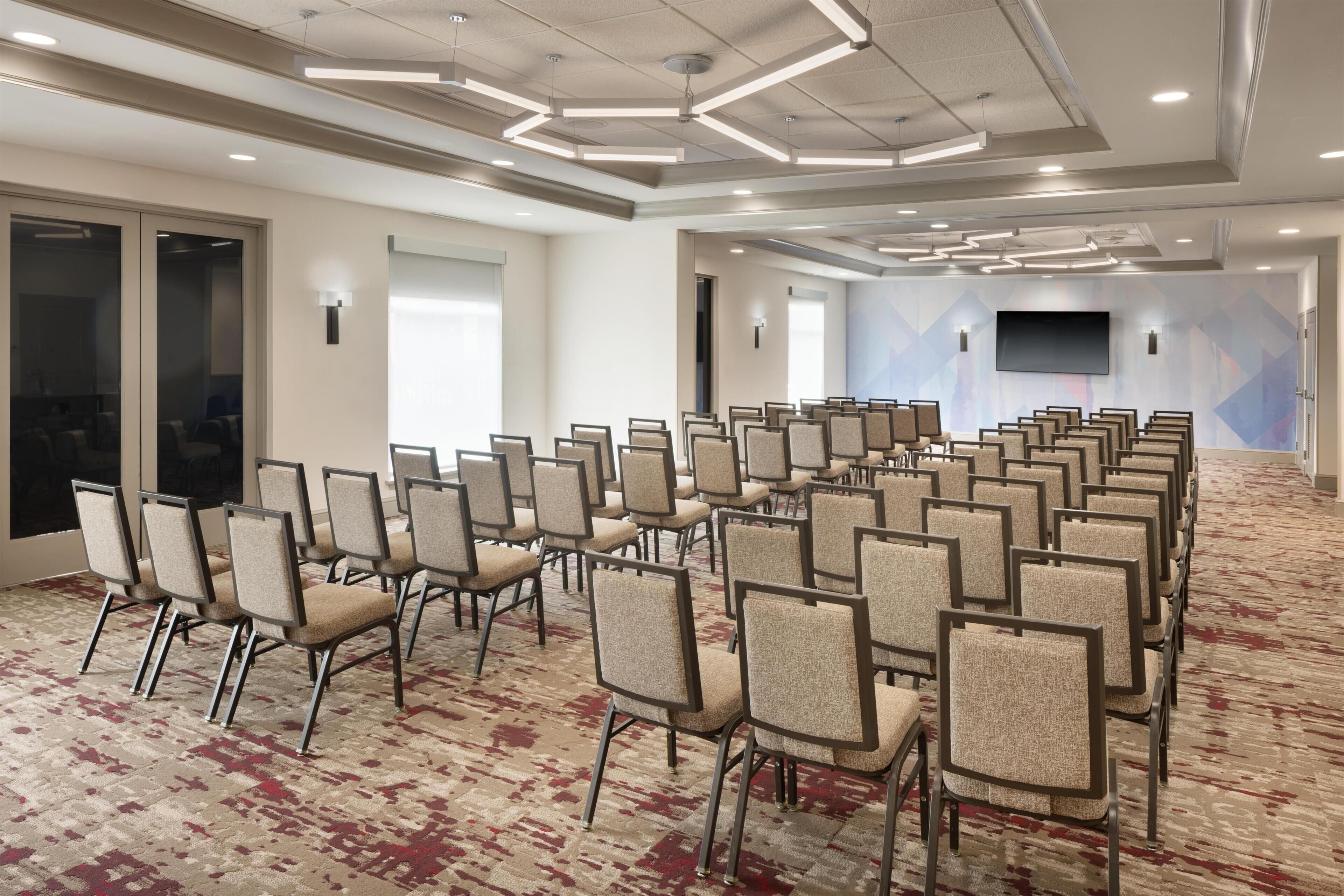A conference room with rows of chairs, a patterned carpet, and a flat-screen TV on the wall.