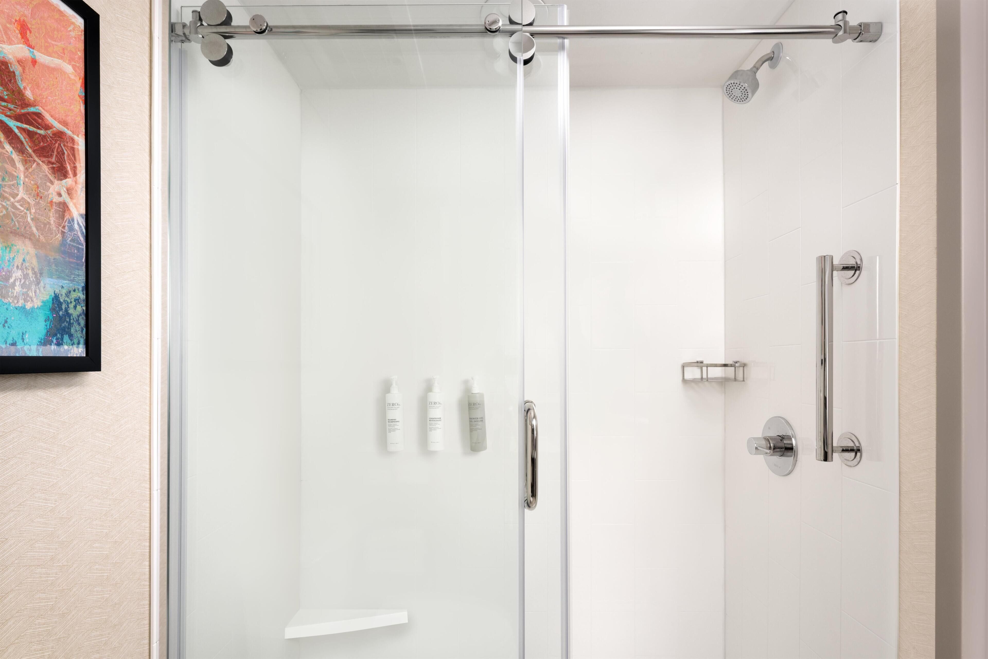 Een douchecabine met glazen wanden, een verchroomde douchekop, een handdouche en een bedieningspaneel met drie flessen shampoo.