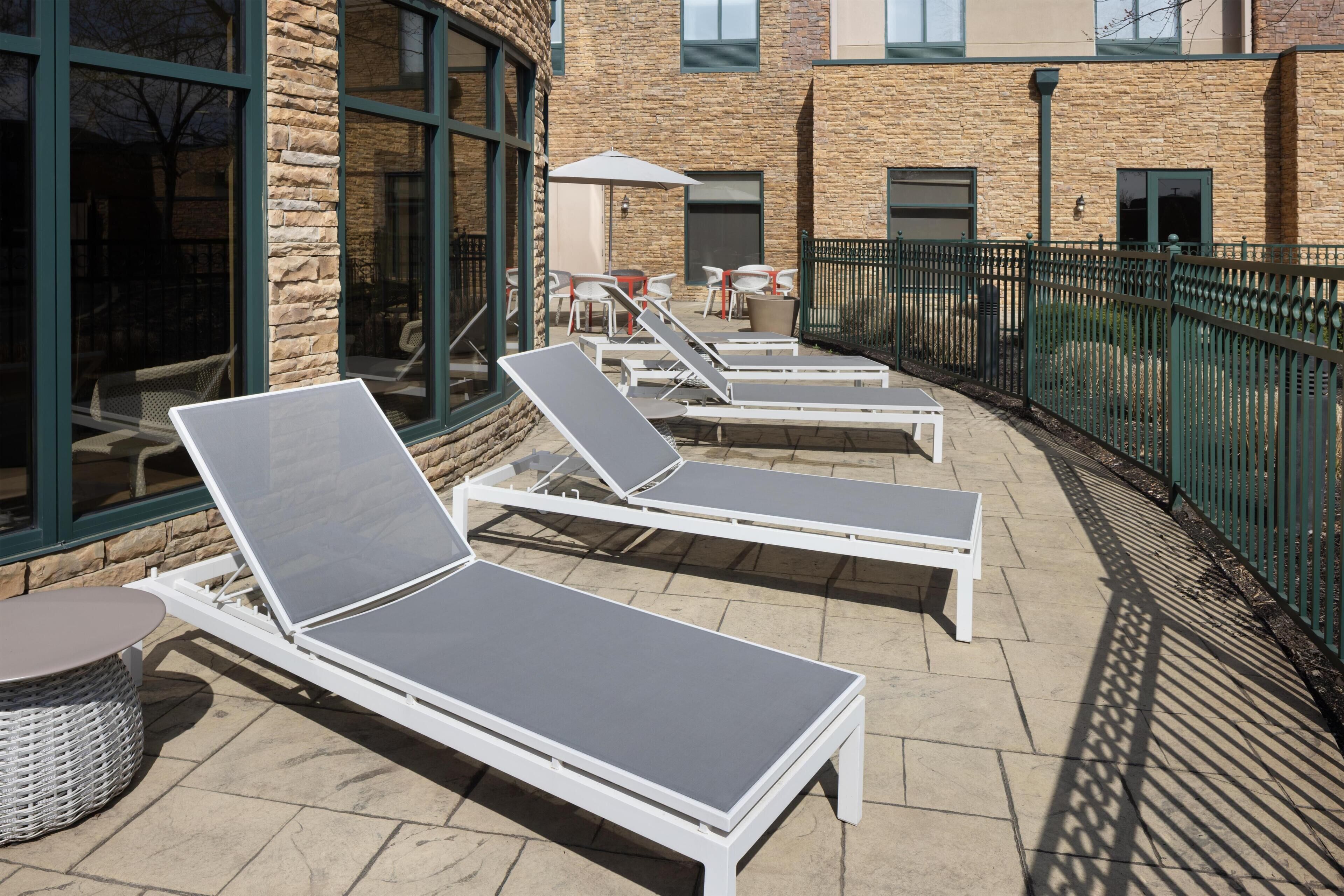 Outdoor lounge chairs and tables arranged on a patio with stone flooring, a stone building in the background, and a green metal fence.