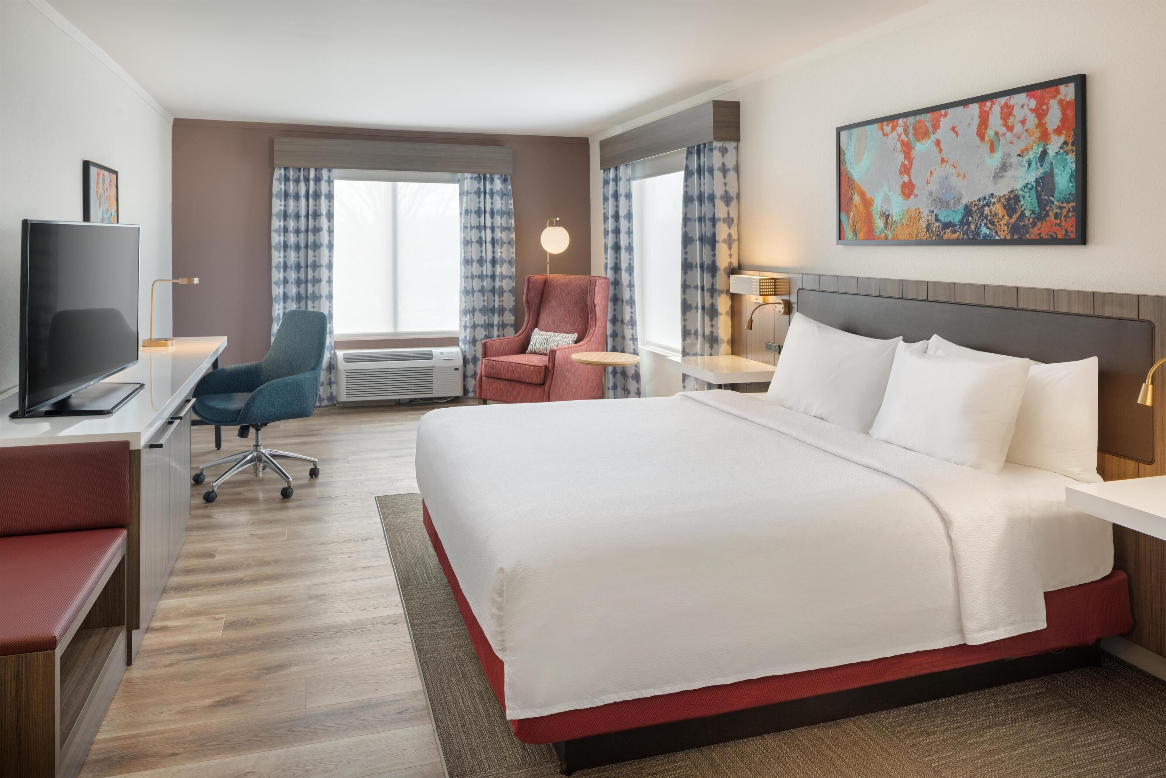 Een hotelkamer met een groot bed, een bureau met een televisie, een stoel en een raam met gordijnen.