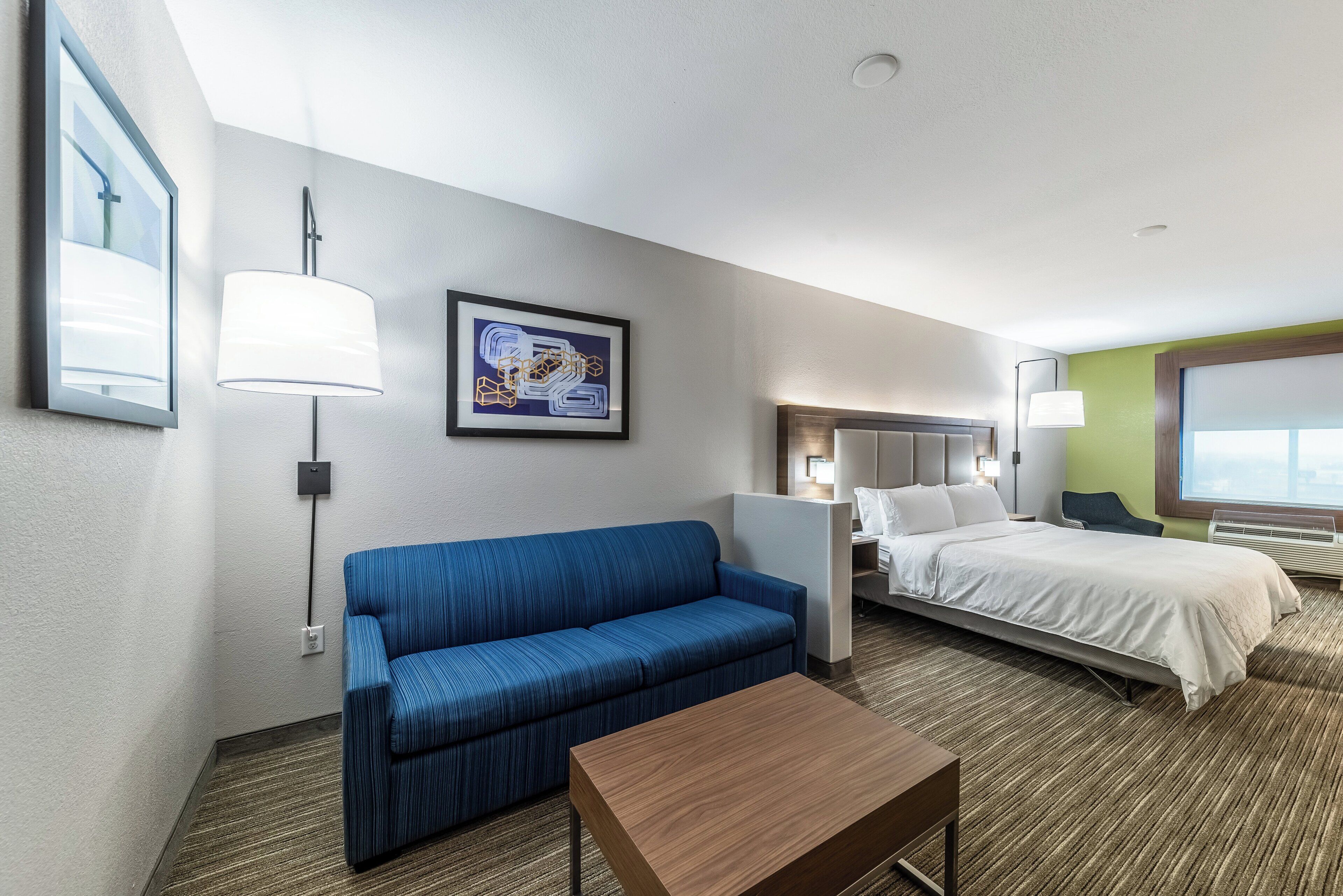 Une chambre d’hôtel avec un canapé bleu, une table en bois, un lit avec une literie blanche, une tête de lit et un tableau encadré au mur.