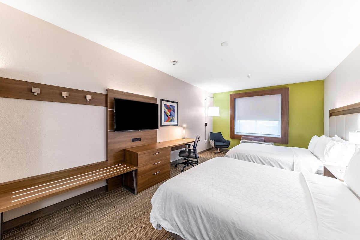 A hotel room with a wooden desk, a flat-screen TV, two beds, a chair, and a window.