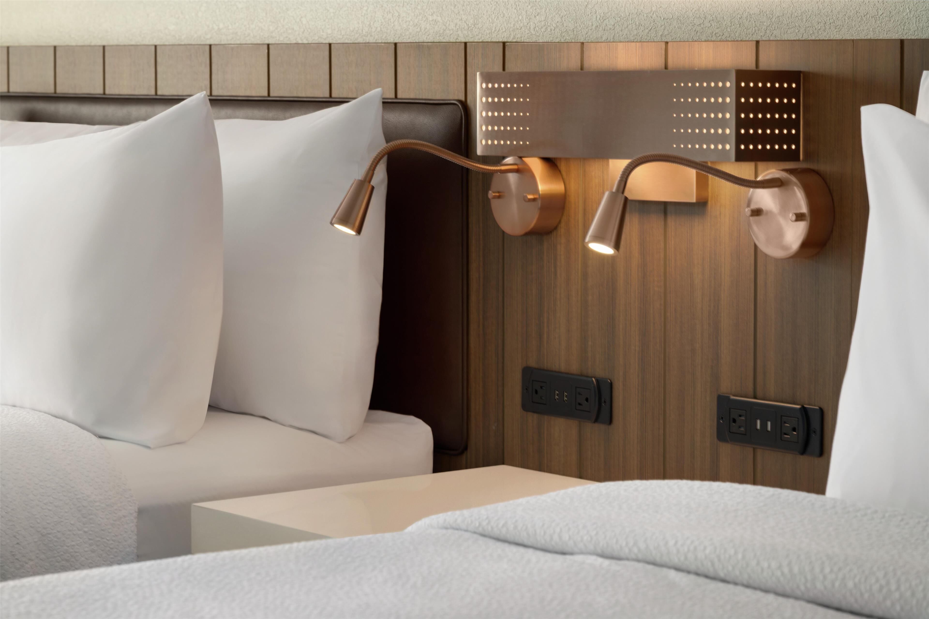 A modern hotel room with a wooden headboard, a bedside table, and a wall-mounted lamp.