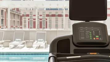 Ein stationäres Fahrrad mit Digitalanzeige, ein Liegebereich am Pool und Blick auf die Stadt.