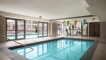 Une piscine intérieure avec des parois en verre transparent, offrant une vue sur le paysage urbain, et dotée d’un éclairage moderne suspendu.