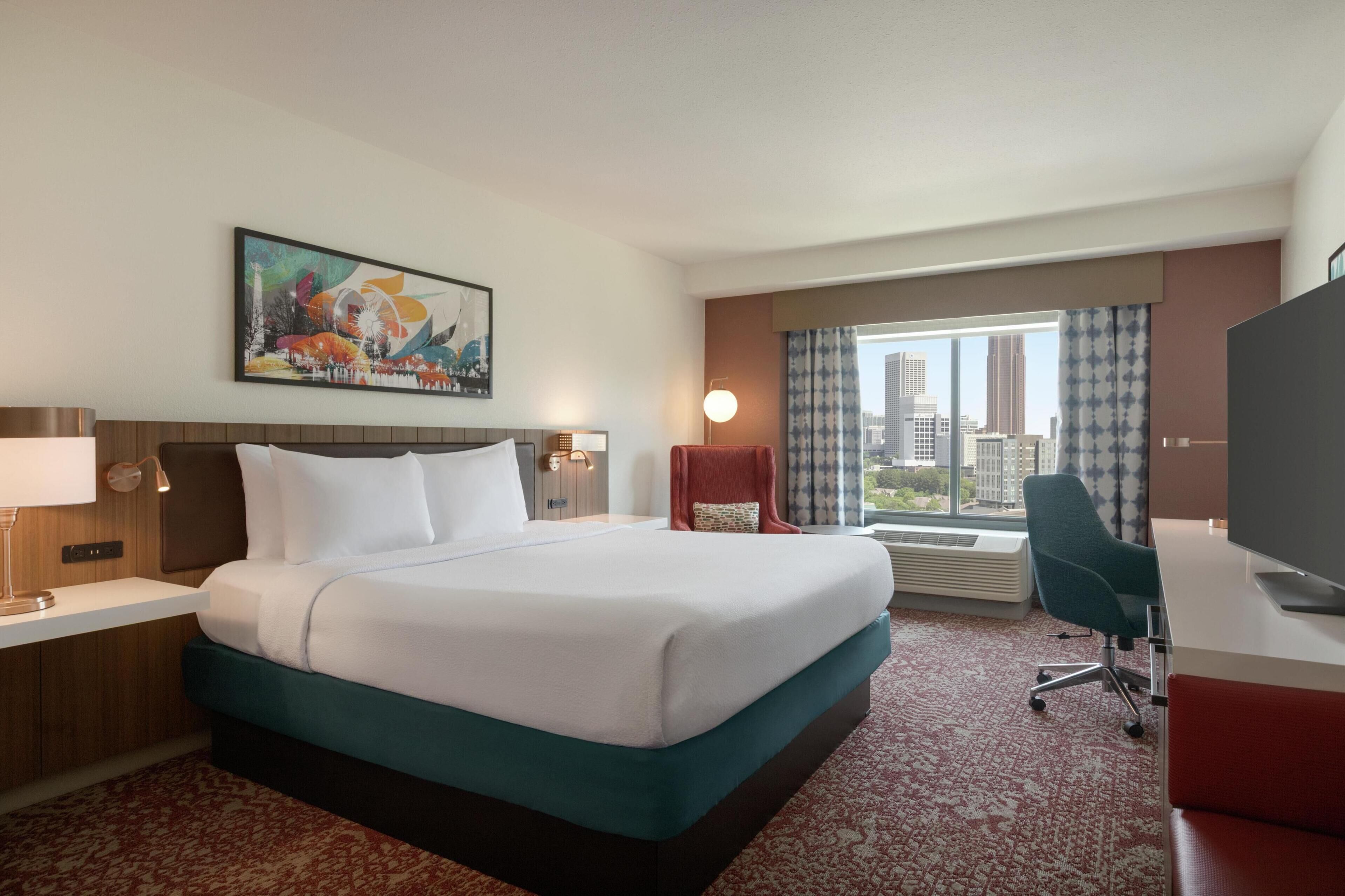 A hotel room with a large bed, a desk with a computer, a chair, a TV, a window with curtains, and a painting on the wall.