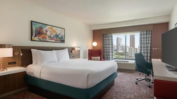 A hotel room with a large bed, a desk with a computer, a chair, a TV, a window with curtains, and a painting on the wall.