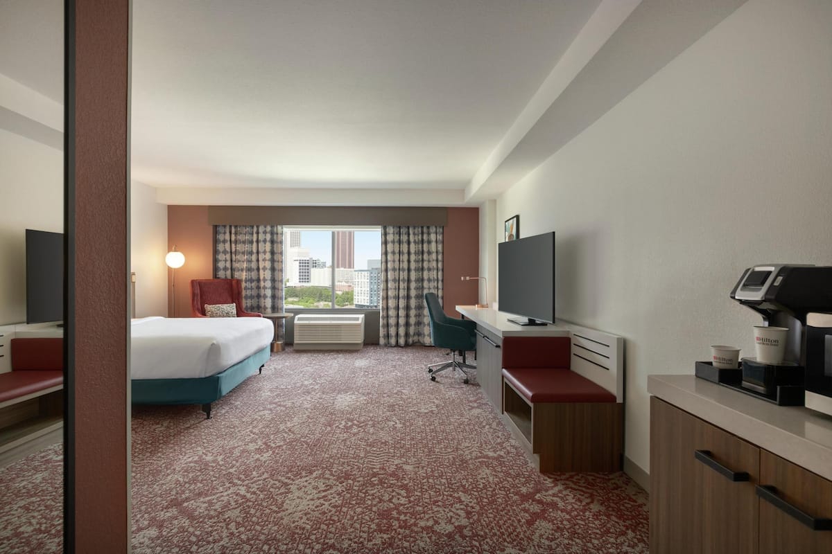 Habitación de hotel con cama, escritorio, silla, televisión y vista a la ciudad.