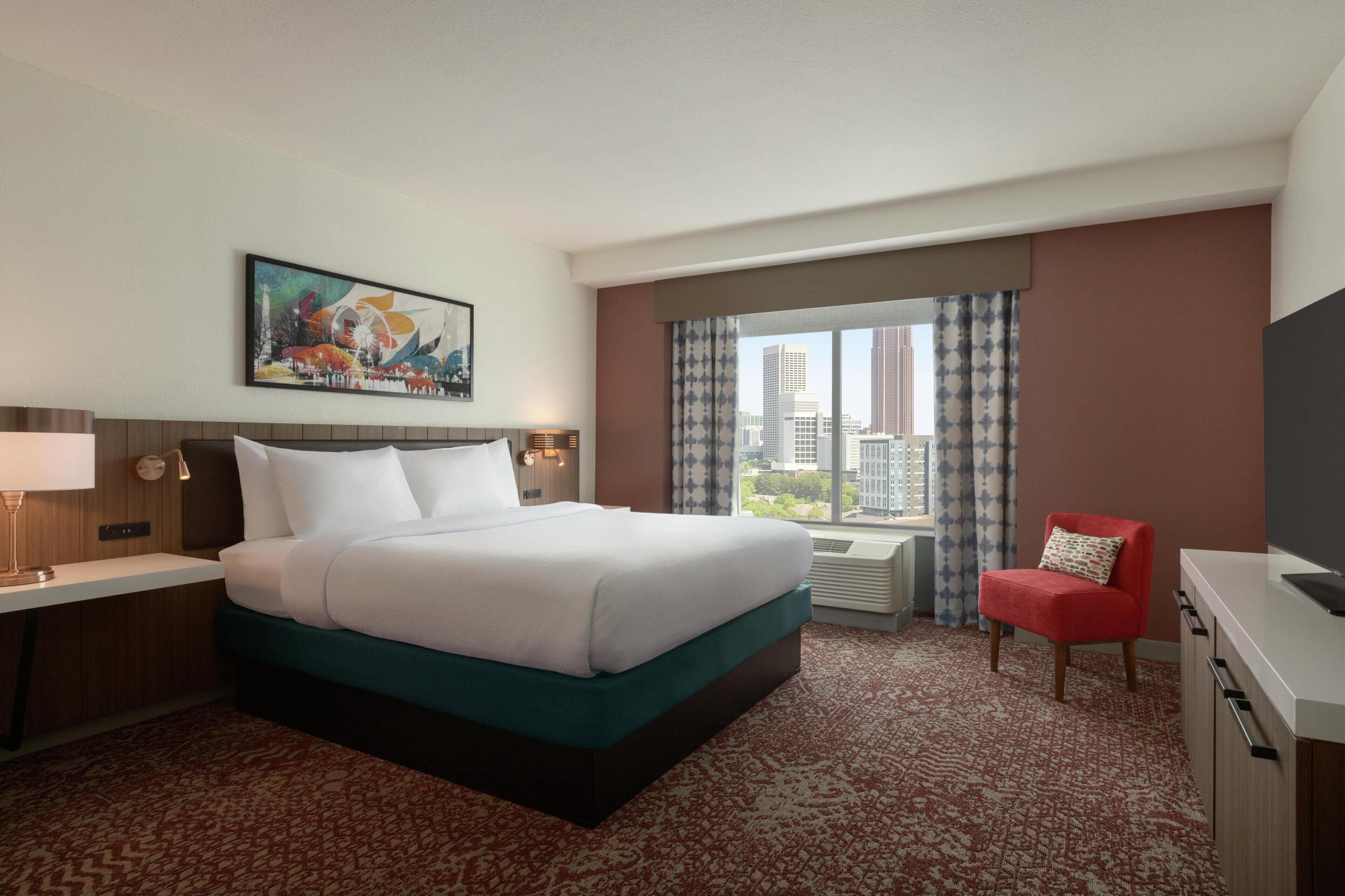 A hotel room with a large bed, a desk, a chair, and a window with a city view.