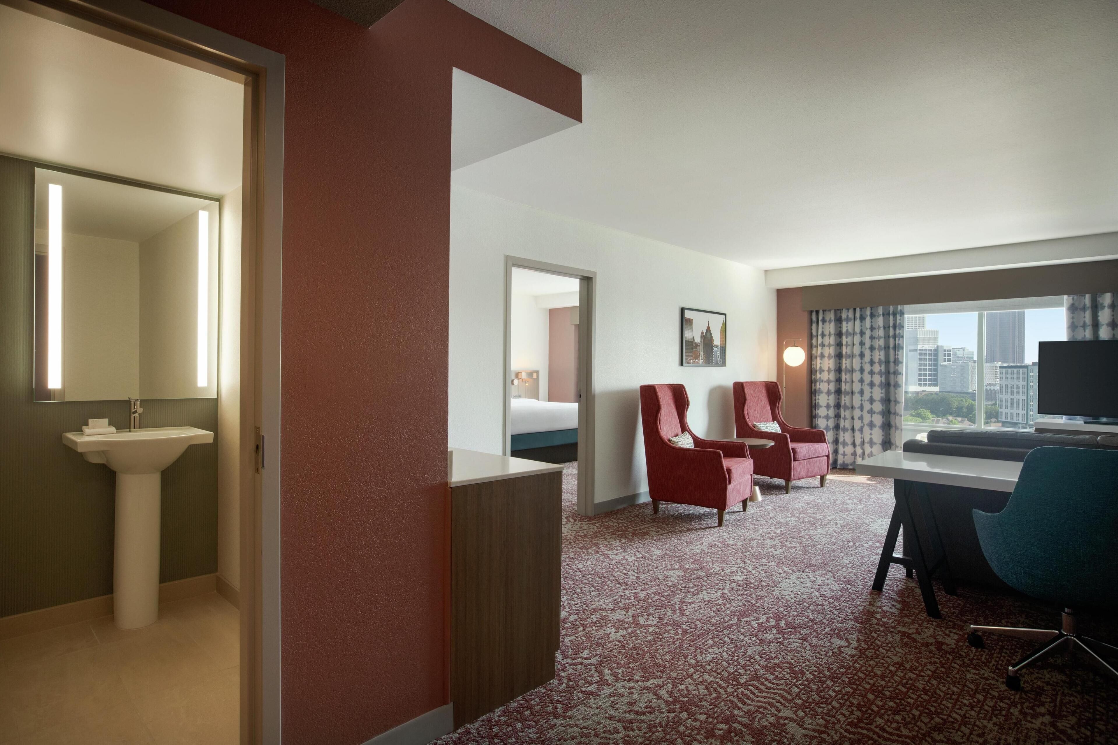 A hotel room with a bed, two red armchairs, a desk with a chair, a large window with a city view, and a bathroom with a sink and mirror.