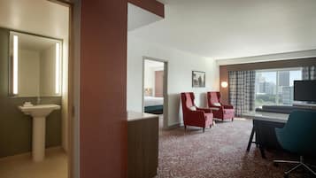 A hotel room with a bed, two red armchairs, a desk with a chair, a large window with a city view, and a bathroom with a sink and mirror.