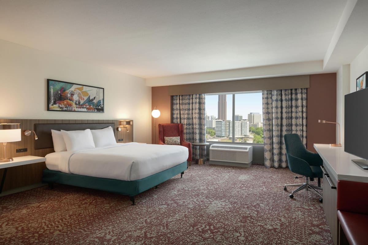 Una habitación de hotel con una cama grande, un escritorio, una silla, un ventanal con vista a la ciudad y un cuadro en la pared.